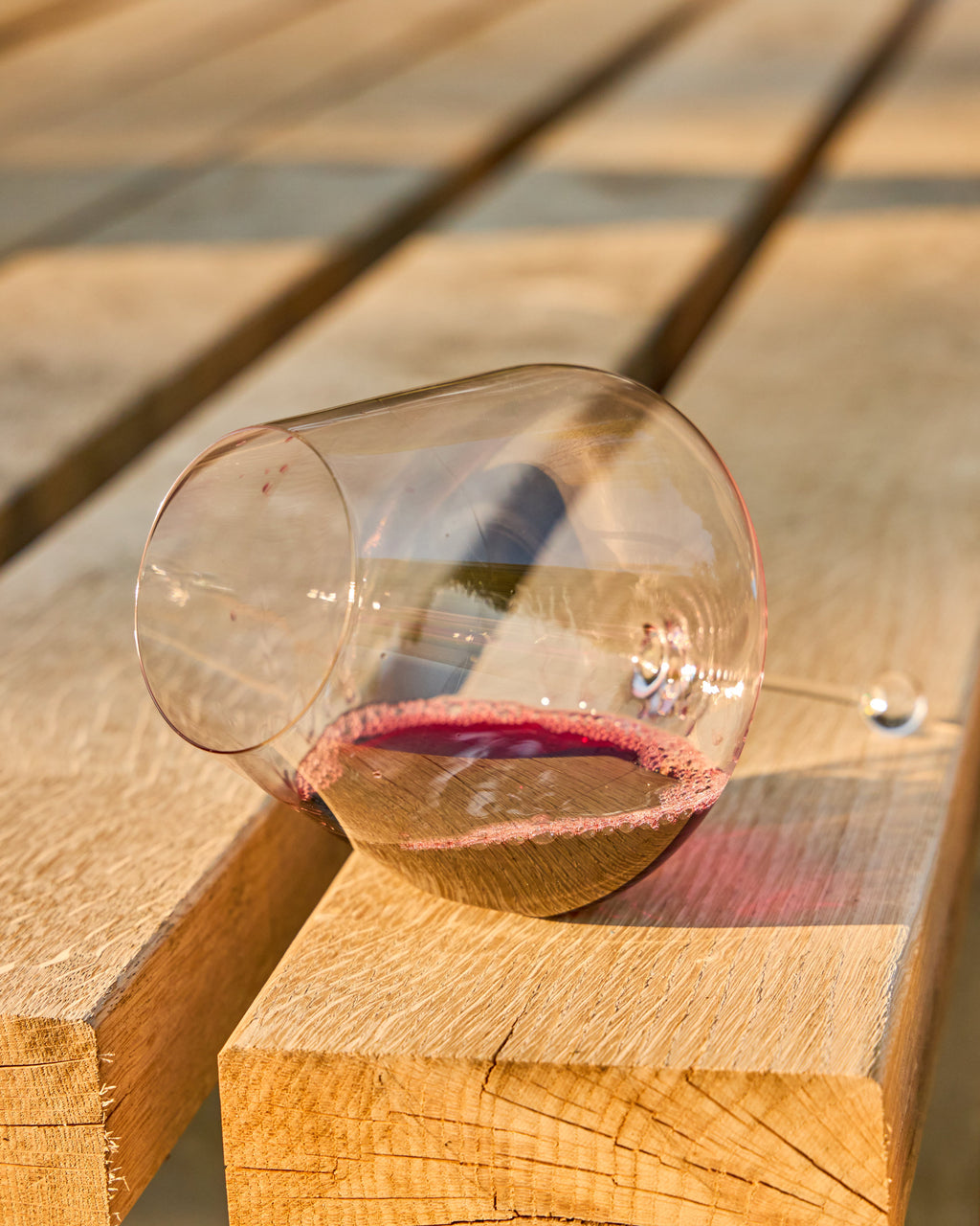 A clear stemmed Zalto Gravitas Omega wine glass lying on its side on wooden planks, with a small amount of red wine residue visible inside and warm sunlight casting shadows across the scene.