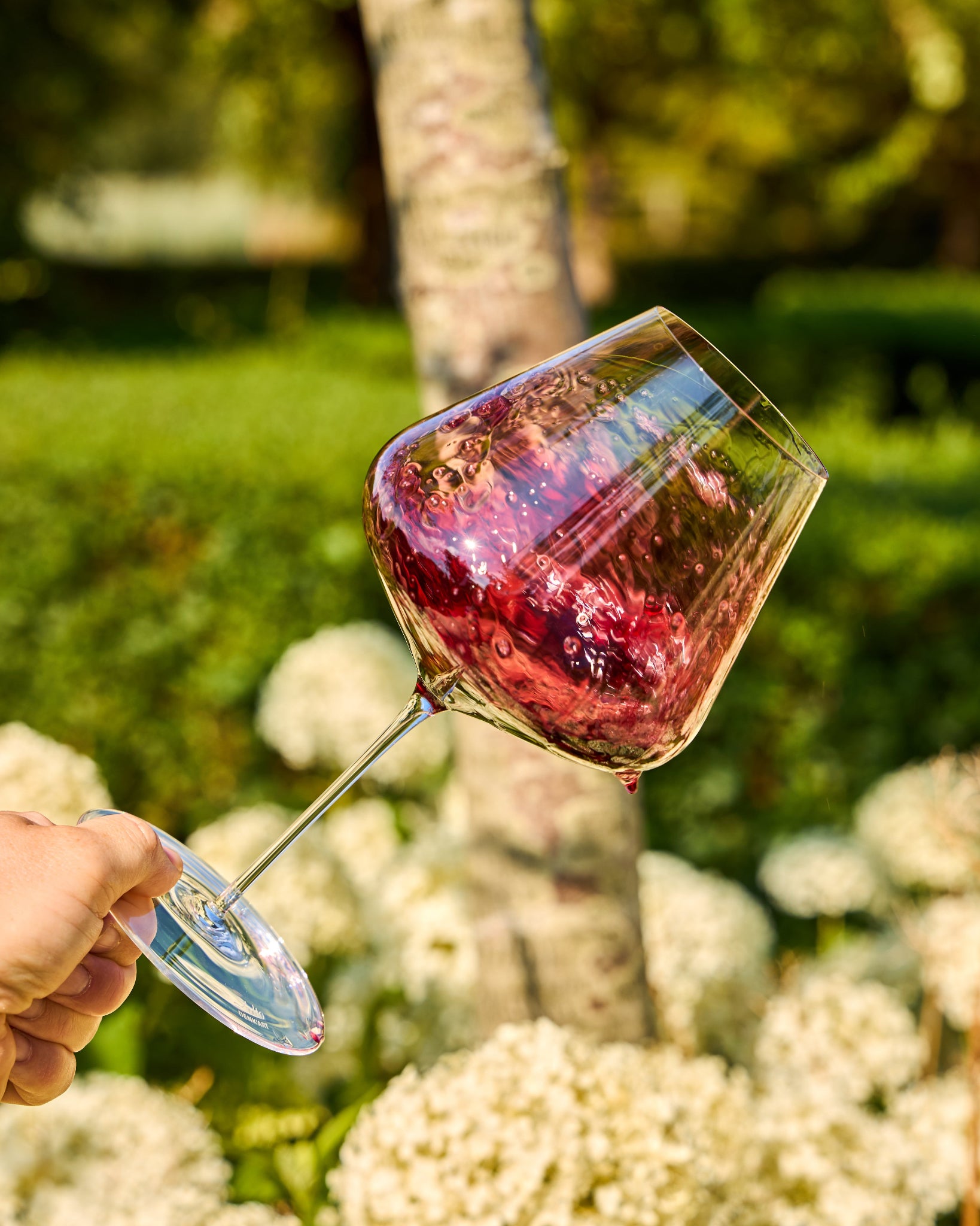 A hand holds a clear Zalto Burgundy wine glass containing deep pink liquid in an outdoor garden setting with a tree trunk and white flowers in the background.